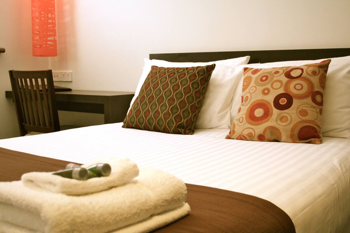 A neatly made bed with patterned pillows, a wooden chair, and a folded towel with bottles on a wooden nightstand.