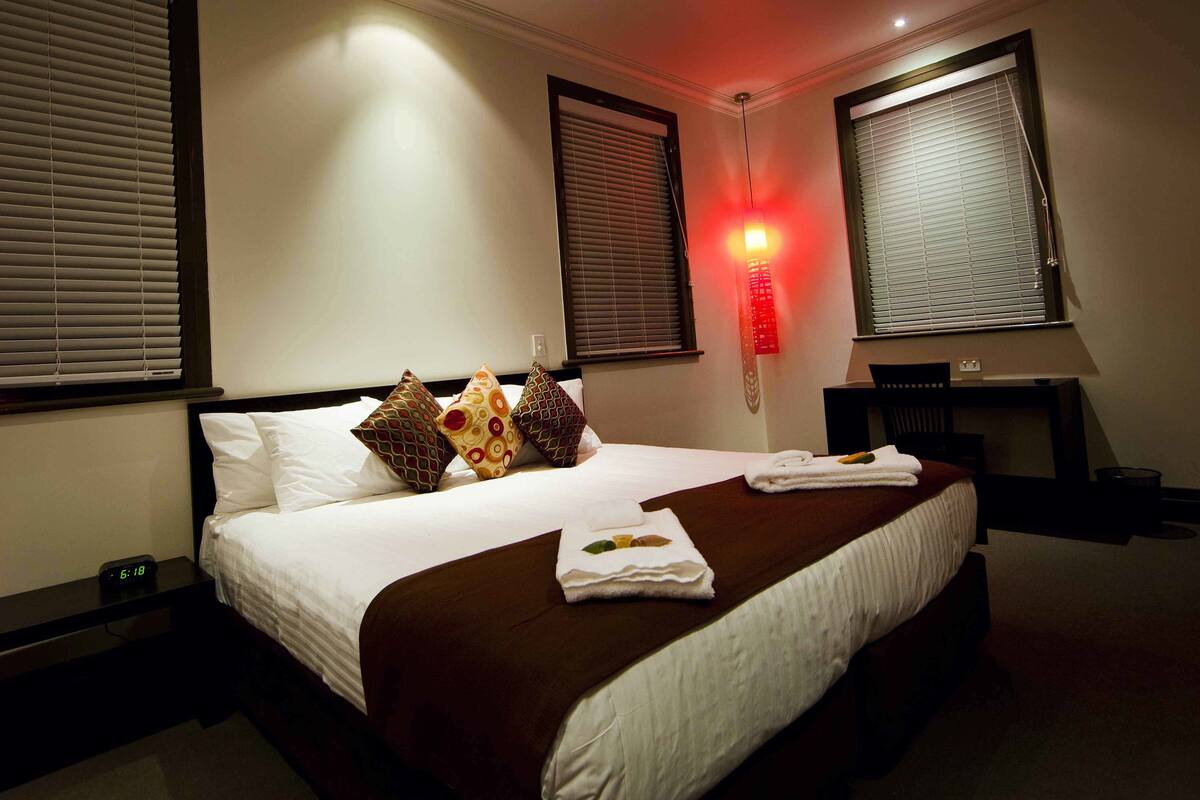 A hotel room with a neatly made bed, a desk, and a red wall light.