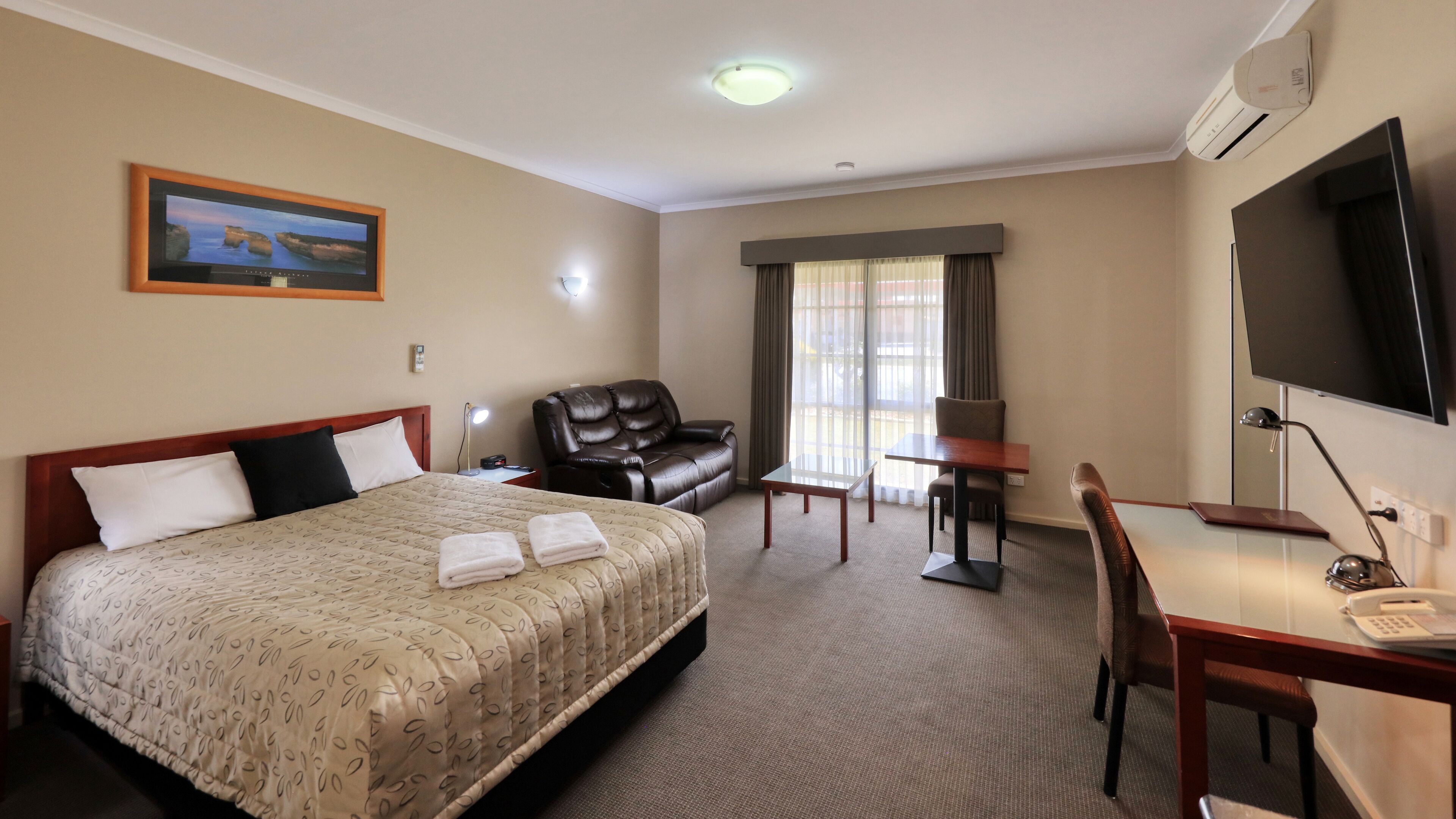 Habitación de hotel con una cama grande, un escritorio, una silla, un sofá de cuero, un televisor y una ventana con cortinas.