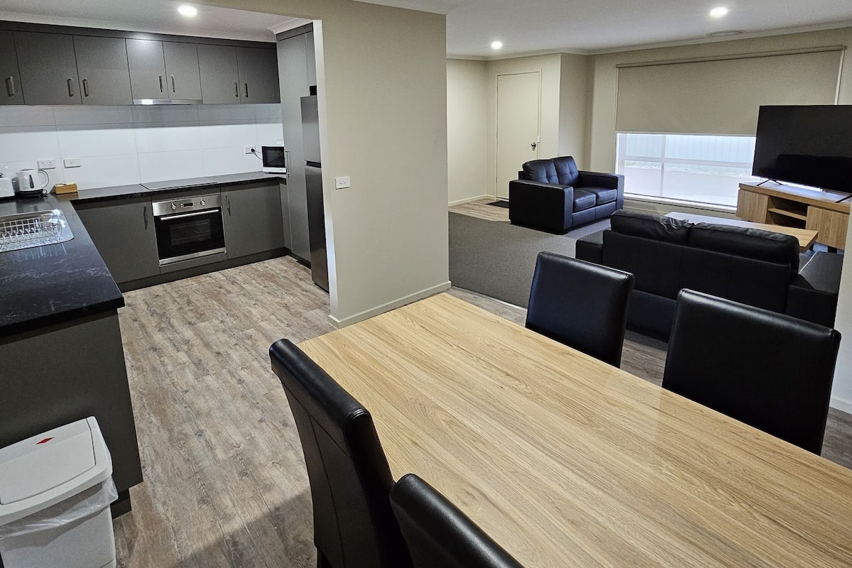 Une cuisine moderne avec des meubles gris, un four à micro-ondes et un évier. On y trouve un coin repas avec une table en bois et des chaises noires. Un canapé et un téléviseur sont visibles au fond.