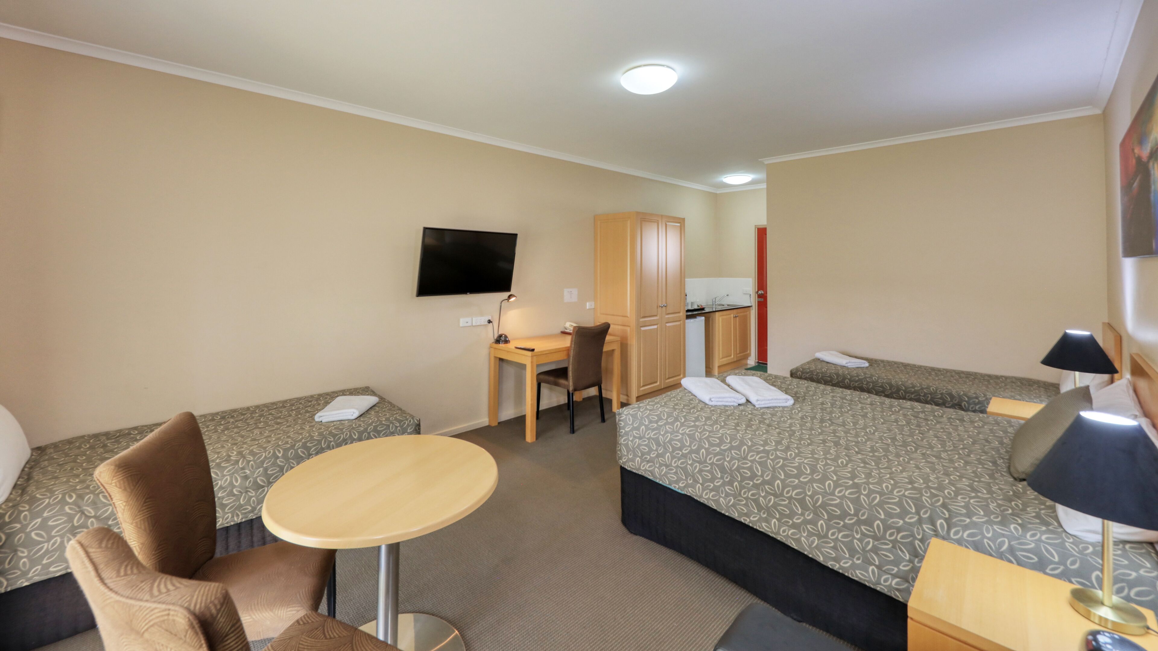 A hotel room with two single beds, a sofa bed, a desk with a chair, a television, and a small kitchenette.
