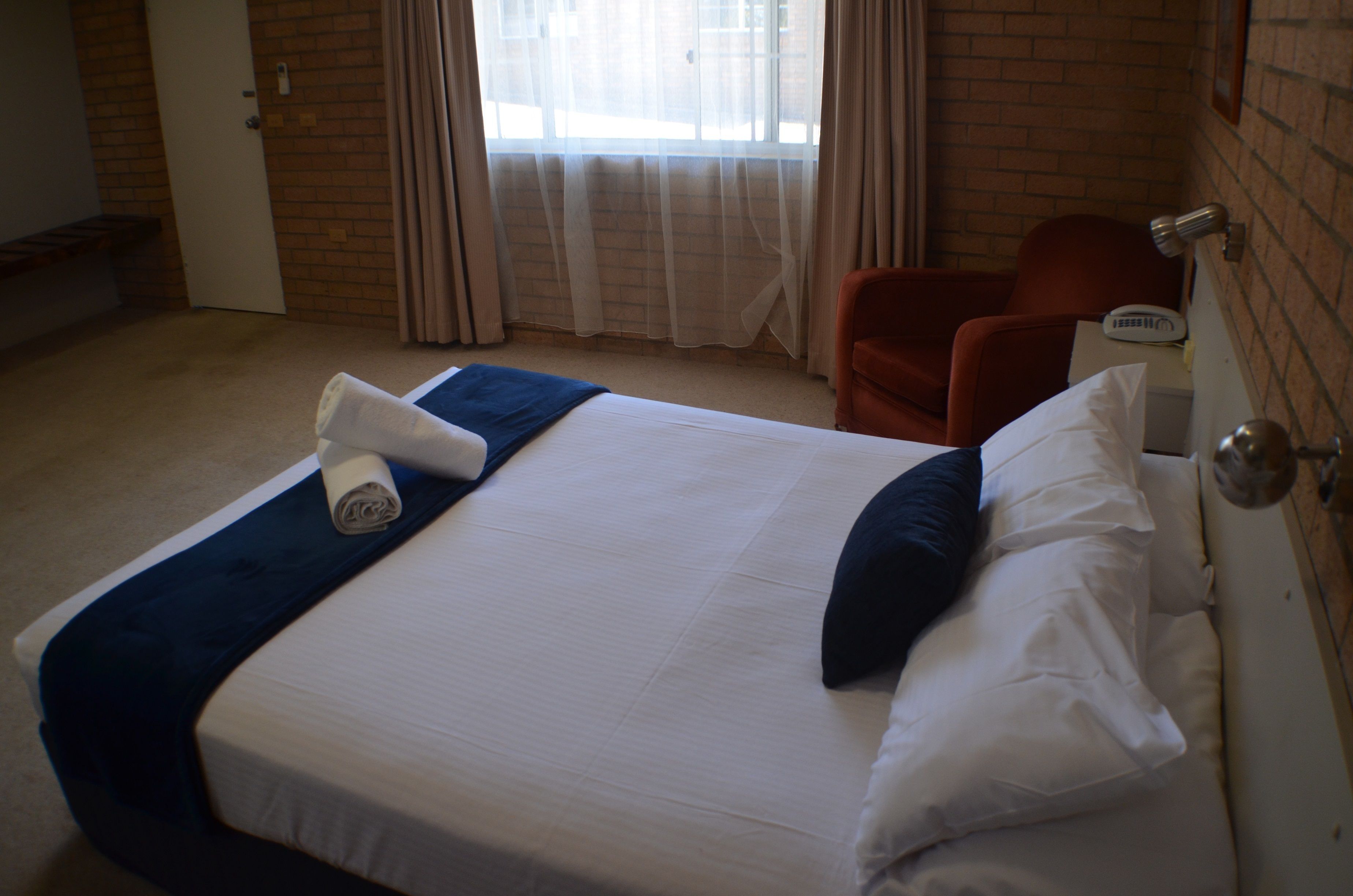 Una cama bien hecha con una manta azul y almohadas, un sillón rojo, una mesita de noche con una lámpara y una ventana con cortinas.