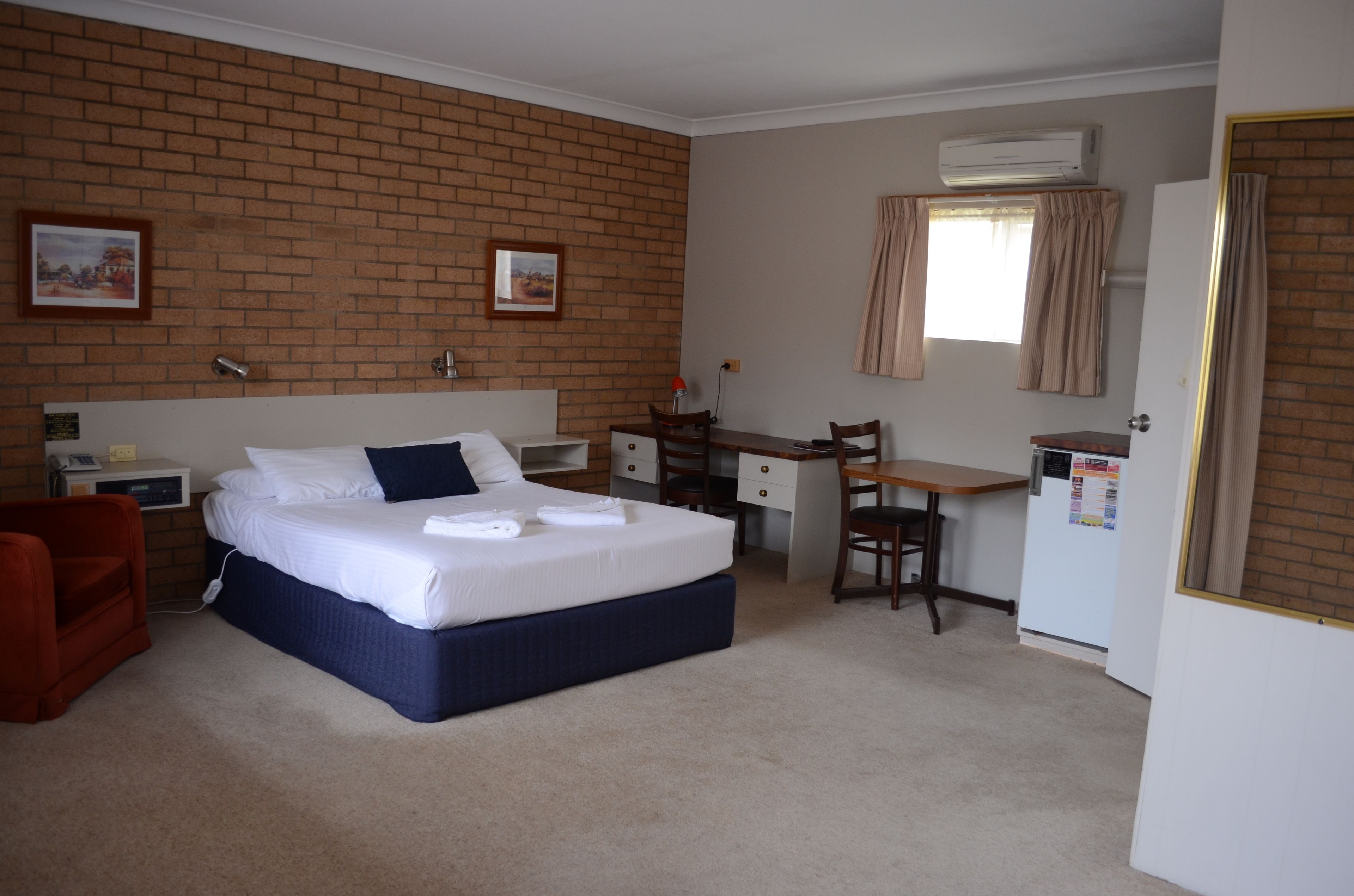 A hotel room with a brick wall, a bed with white bedding, a desk with a chair, a small refrigerator, and an air conditioning unit.