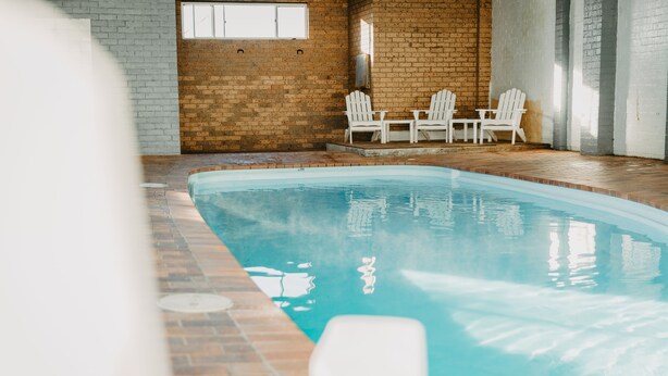 Ein überdachter Swimmingpool mit Backsteinwand und weißen Stühlen.