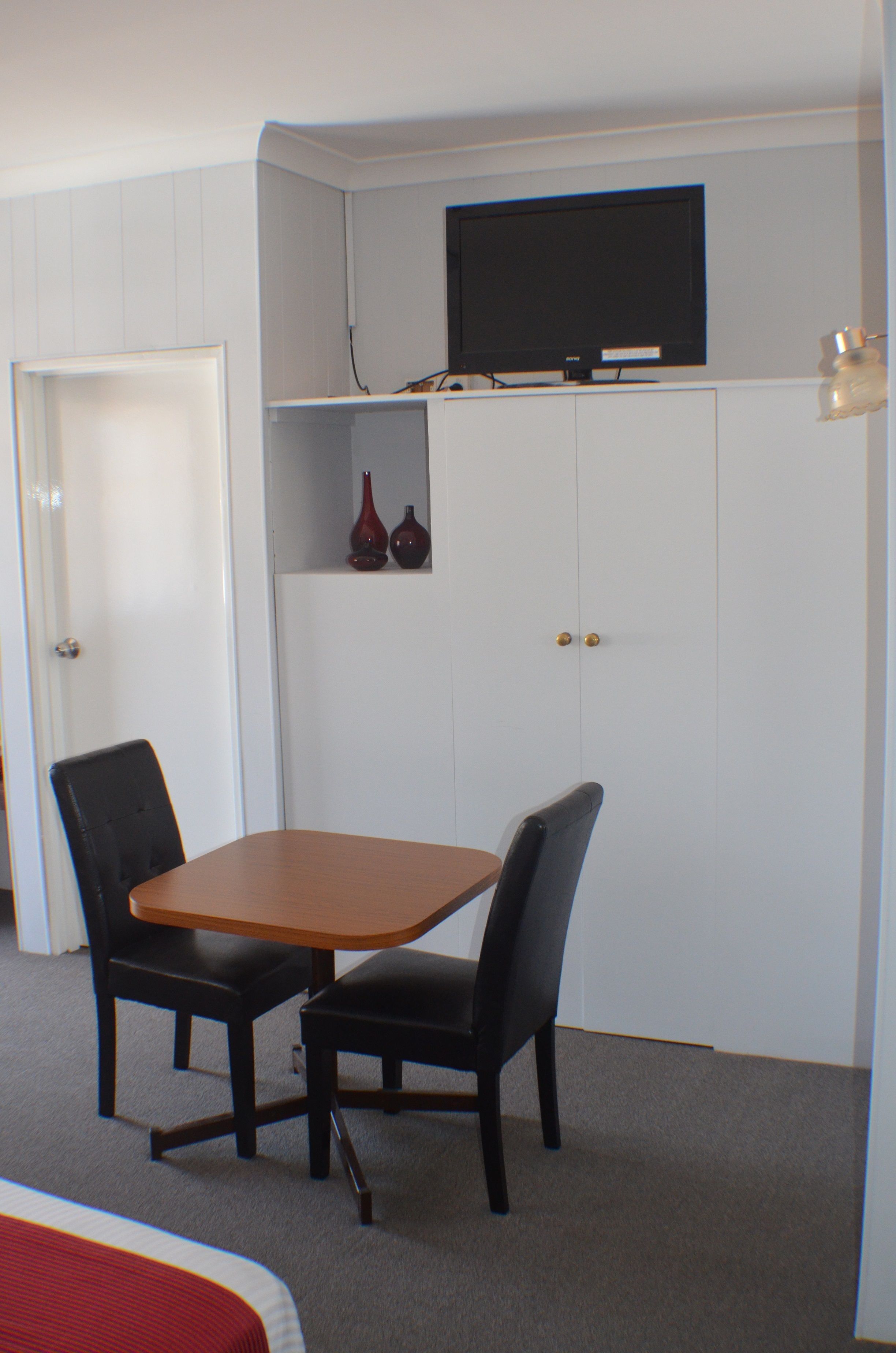A room with a bed, a table with two chairs, a television on a shelf, and a door.