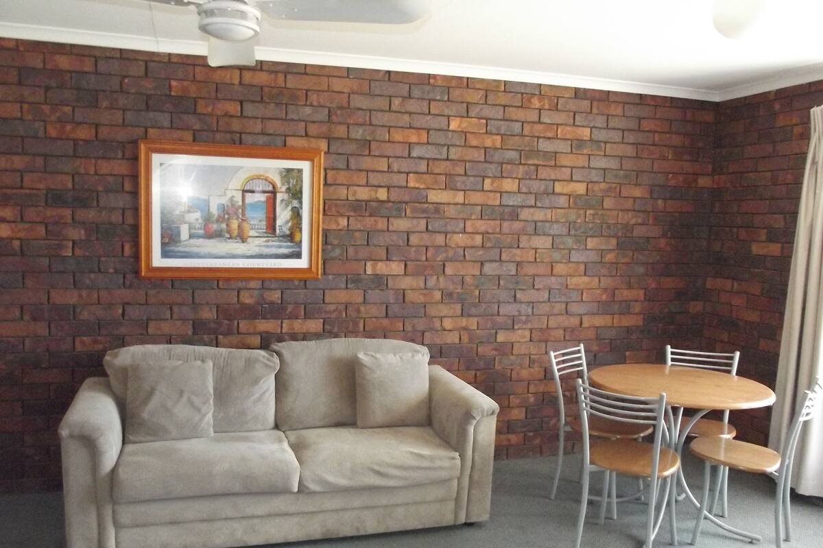 A room with a brick wall, a beige sofa, a small round table with chairs, and a ceiling fan.