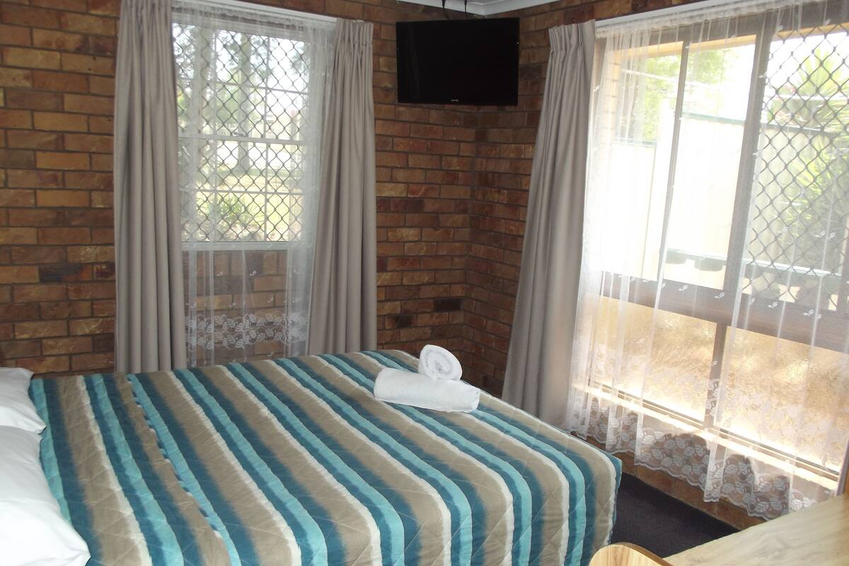 A hotel room with a brick wall, a bed with striped bedding, a TV mounted on the wall, and a window with a view of greenery.