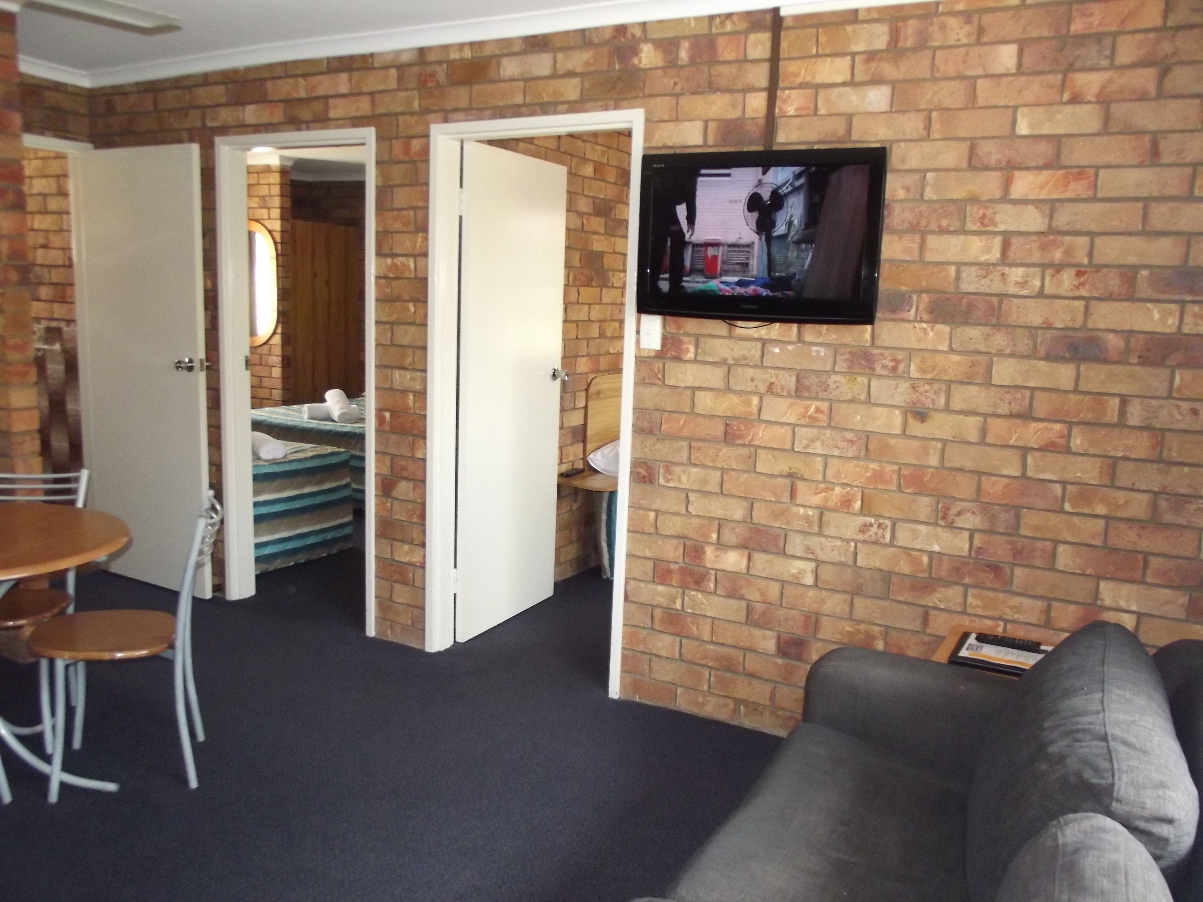 A room with a brick wall, a television mounted on it, a sofa, a dining table with chairs, and a bed in the background.
