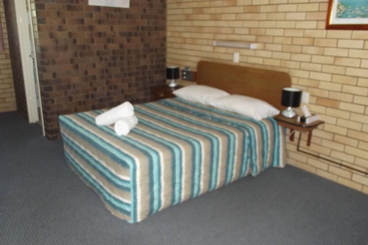 A hotel room with a brick wall, a bed with striped bedding, a wooden headboard, a nightstand with a lamp, and a framed picture on the wall.