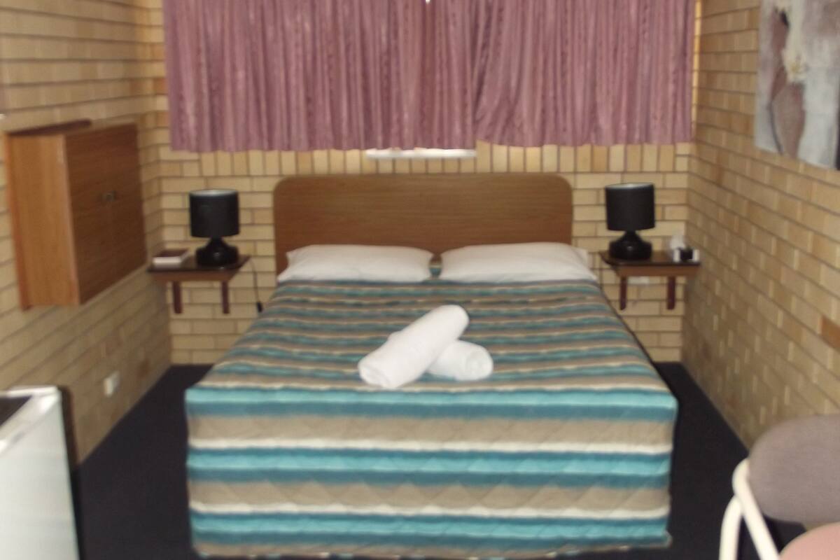 A bedroom with a bed, a ceiling fan, a nightstand with a lamp, a chair, and a painting on the wall.