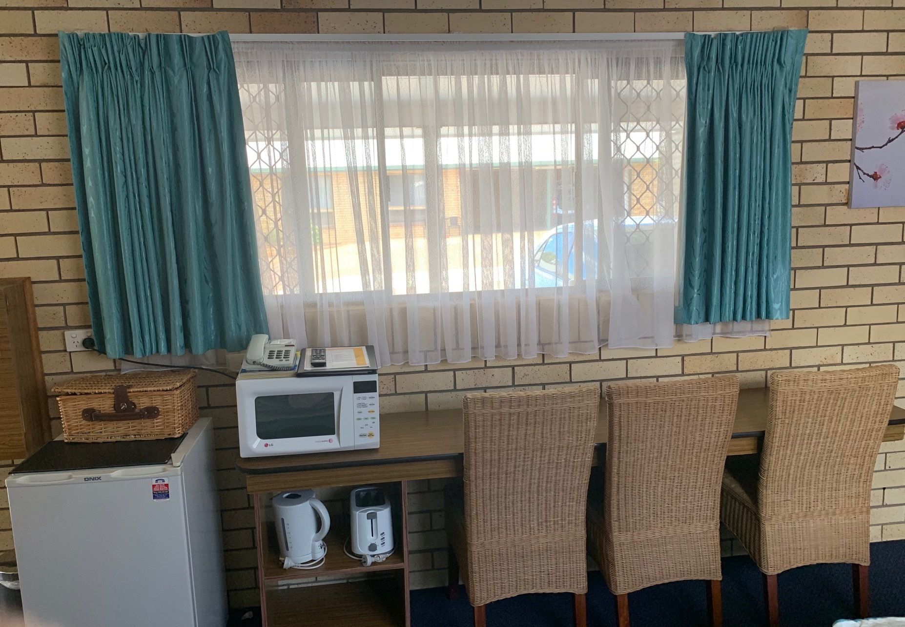 Ein kompakter Raum mit Backsteinwand, einem Fenster mit türkisfarbenen Vorhängen, einer Mikrowelle, einem Toaster und Korbsesseln.