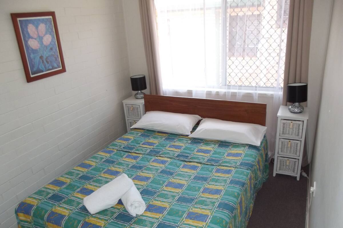A neatly made bed with a colorful quilt, two bedside tables with lamps, a window with curtains, and a framed picture on the wall.