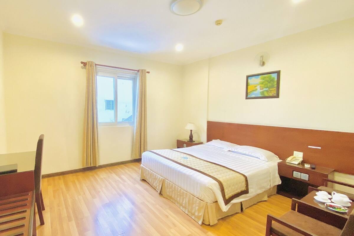A hotel room with a bed, a desk, a chair, a window with curtains, a bedside table with a lamp, and a painting on the wall.