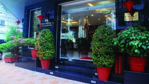 Entrada de un hotel con fachada de vidrio, plantas en macetas rojas y un cartel con el nombre del hotel y su información de contacto.