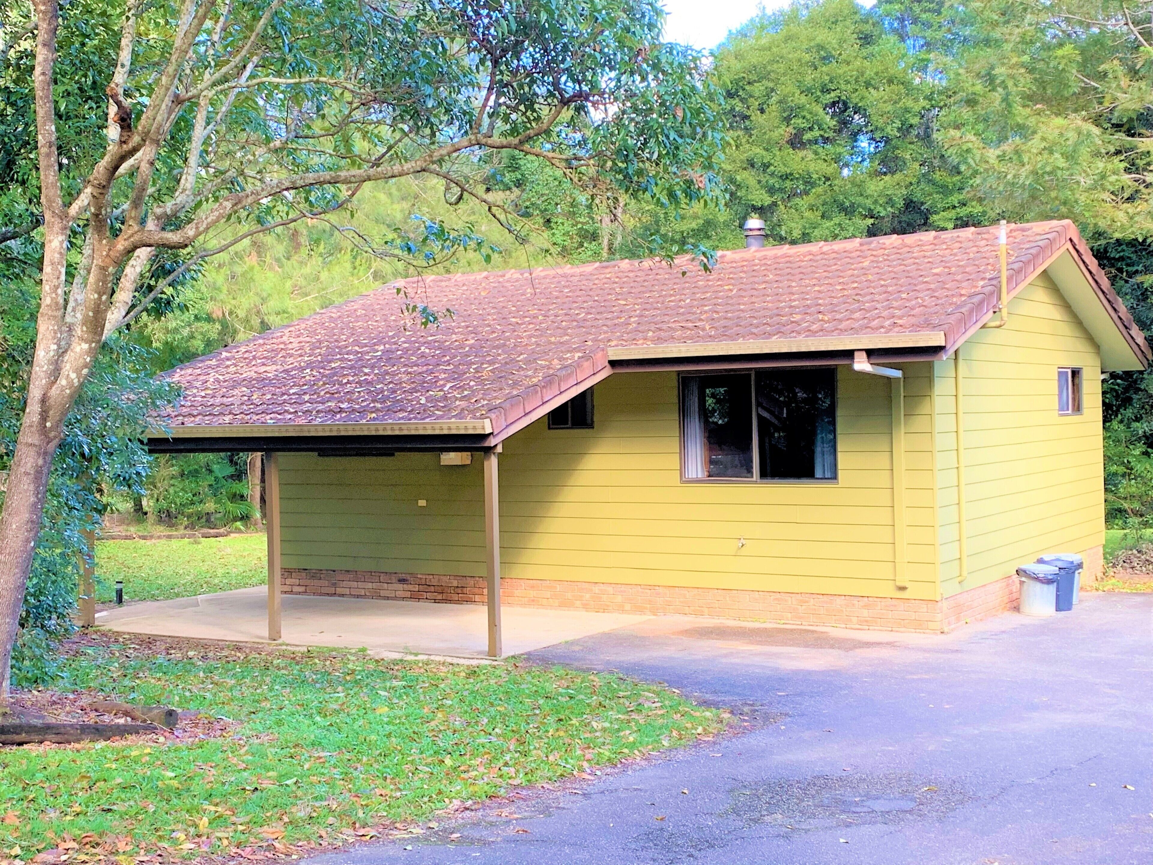 A small yellow building with a red-tiled roof, a covered porch, and a paved driveway.