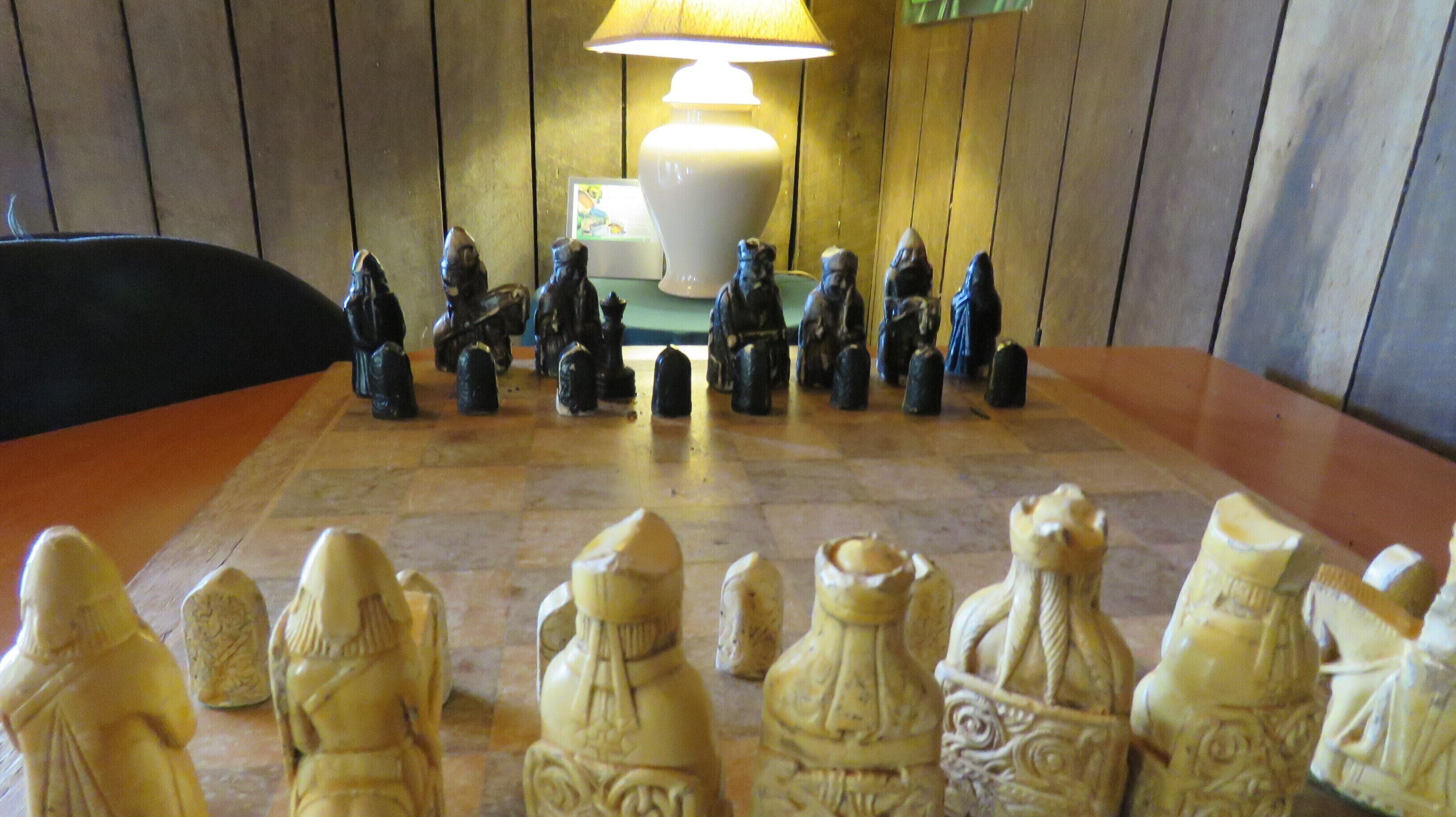 A chessboard with carved figures in a room with wooden paneling and a table lamp.