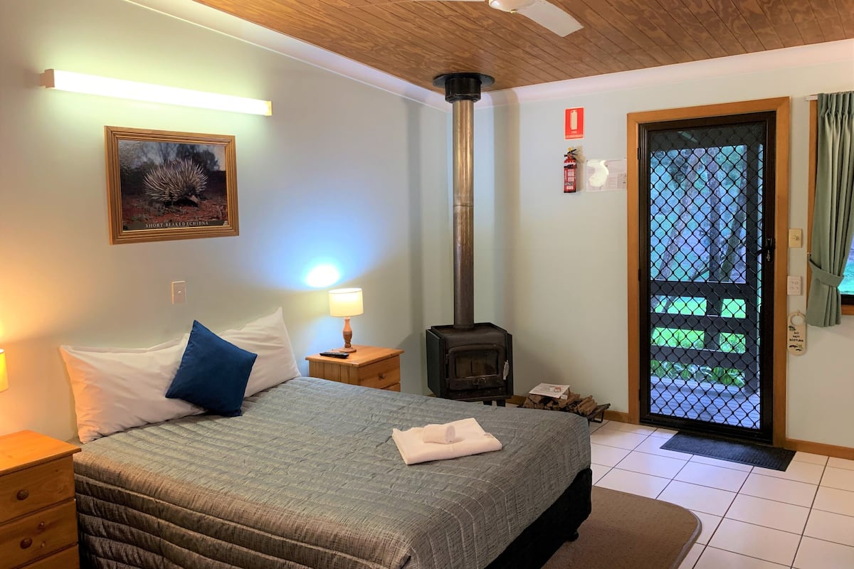 Une chambre à coucher comprenant un lit, une table de chevet, une lampe fixée au mur, un ventilateur de plafond, une cheminée et une porte donnant sur une vue agréable.