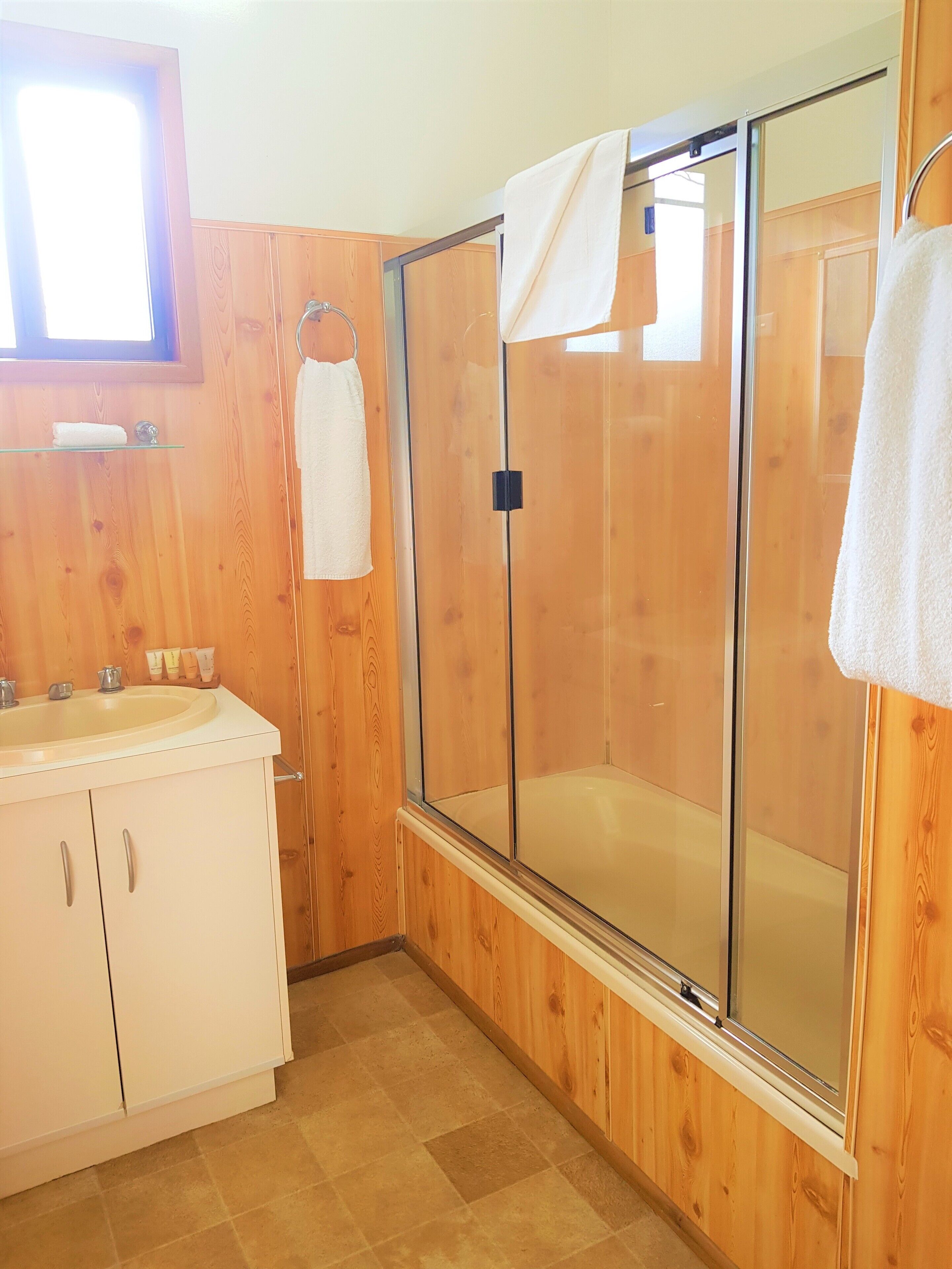 Une salle de bain avec des panneaux de bois, une douche en verre, un lavabo avec des articles de toilette et une fenêtre au-dessus du lavabo.