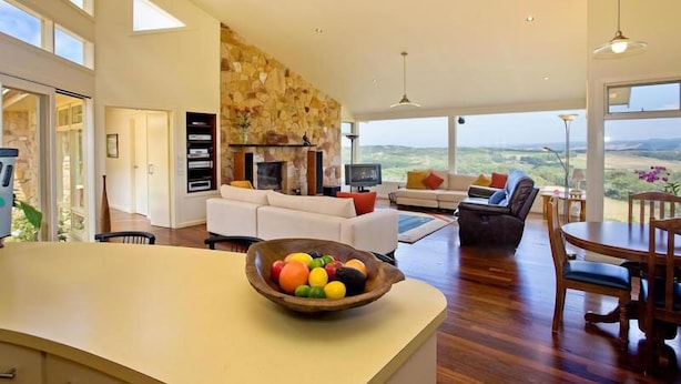 A spacious living area with a stone wall, a fireplace, and a large window offering a view of the countryside. - Cape Otway Cottages (Hordern Vale)