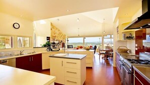 A modern kitchen with a central island, a dining area with a view, and a large window. - Cape Otway Cottages (Hordern Vale)