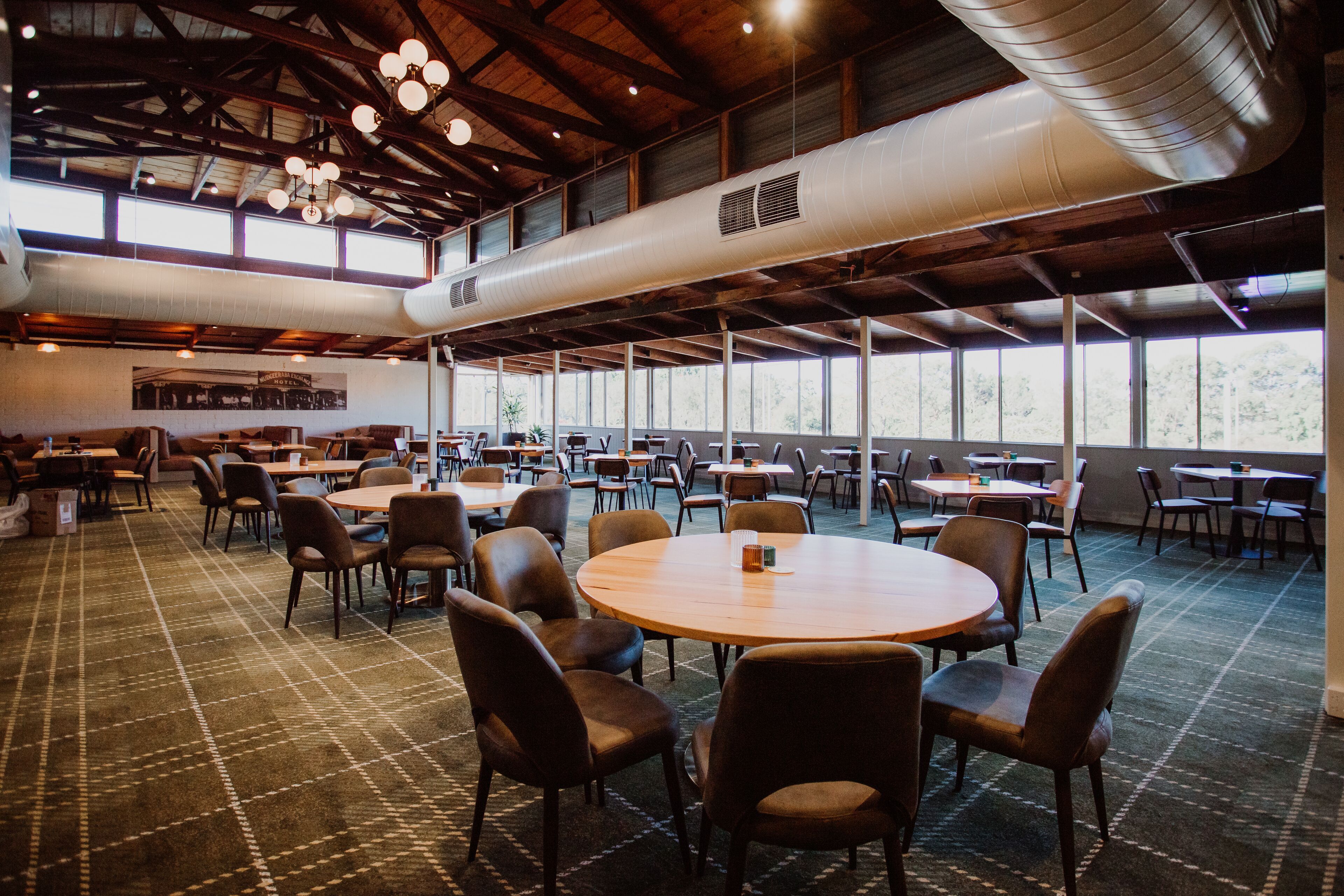 A spacious indoor dining area with round tables and chairs, large windows, and a high ceiling with exposed beams.
