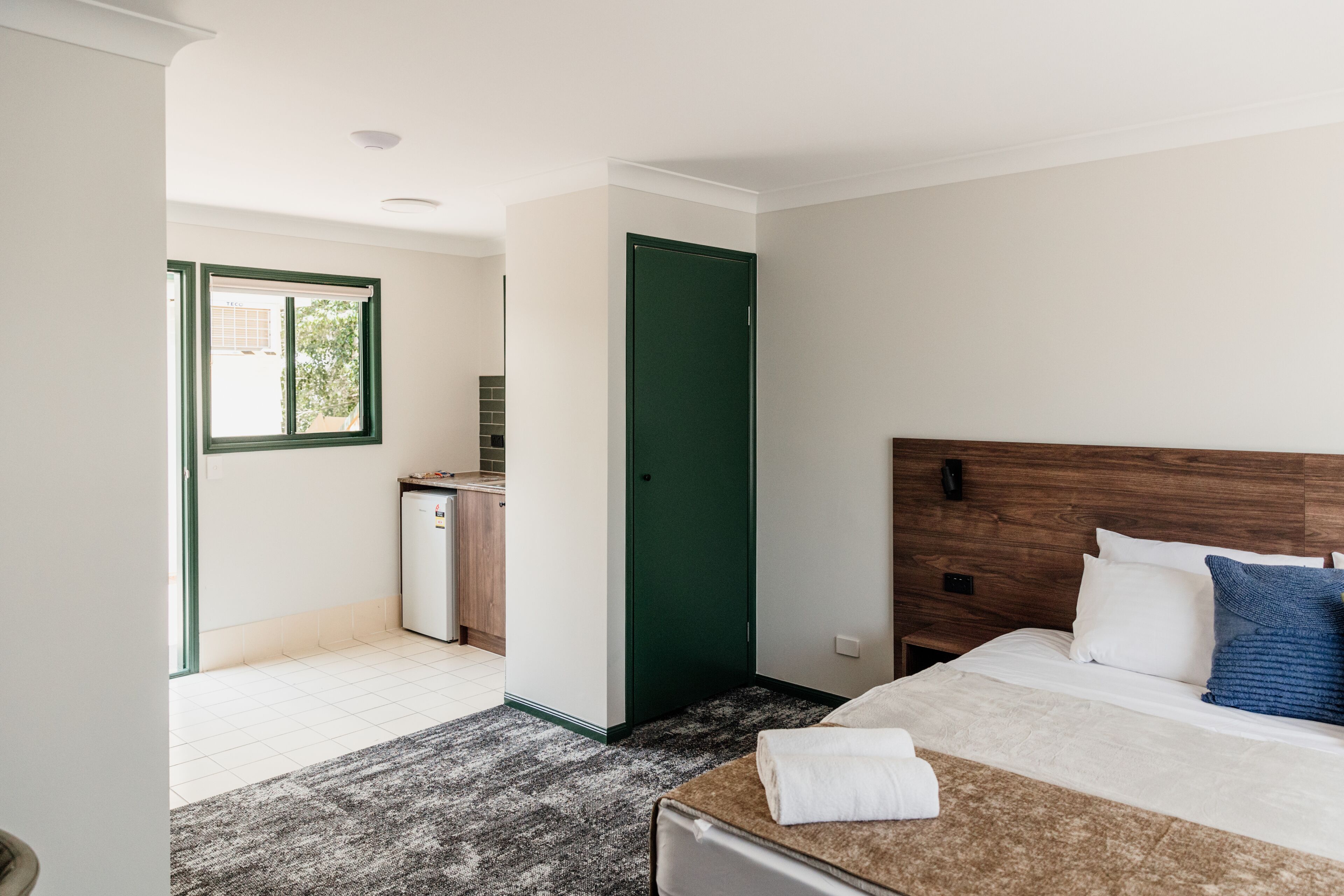 A hotel room with a bed, a small kitchenette, a green door, and a view of the outside.