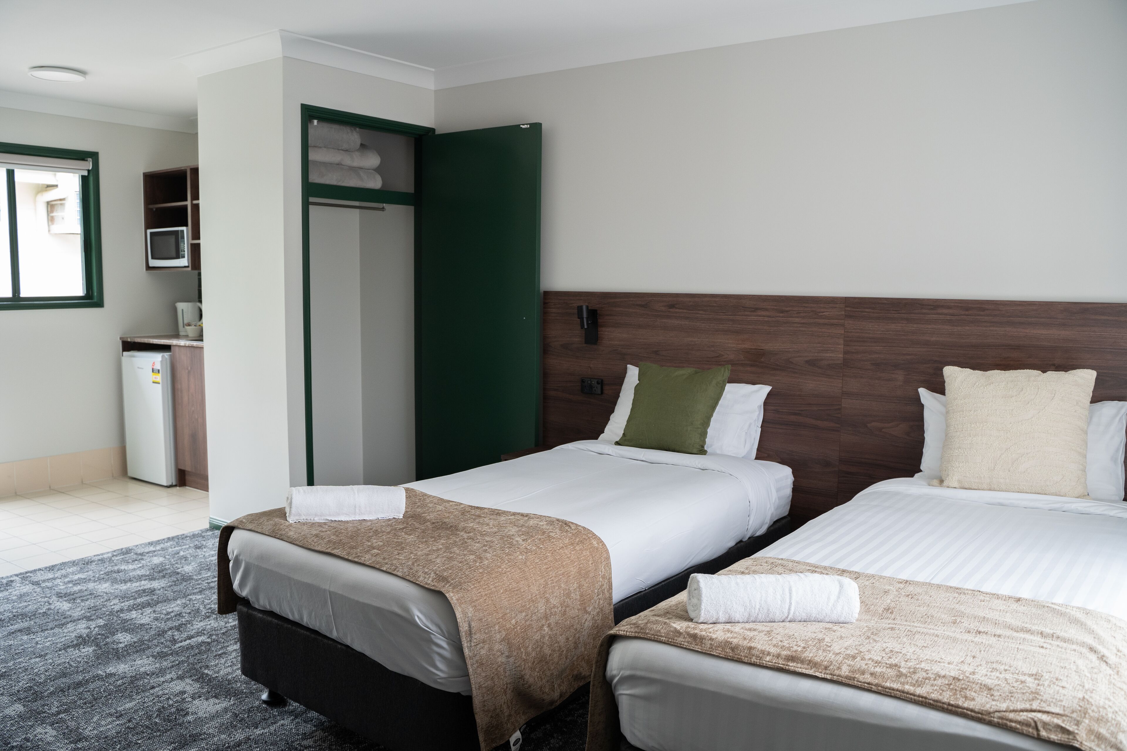 Habitación de hotel con dos camas, un escritorio, un armario y una kitchenette pequeña.
