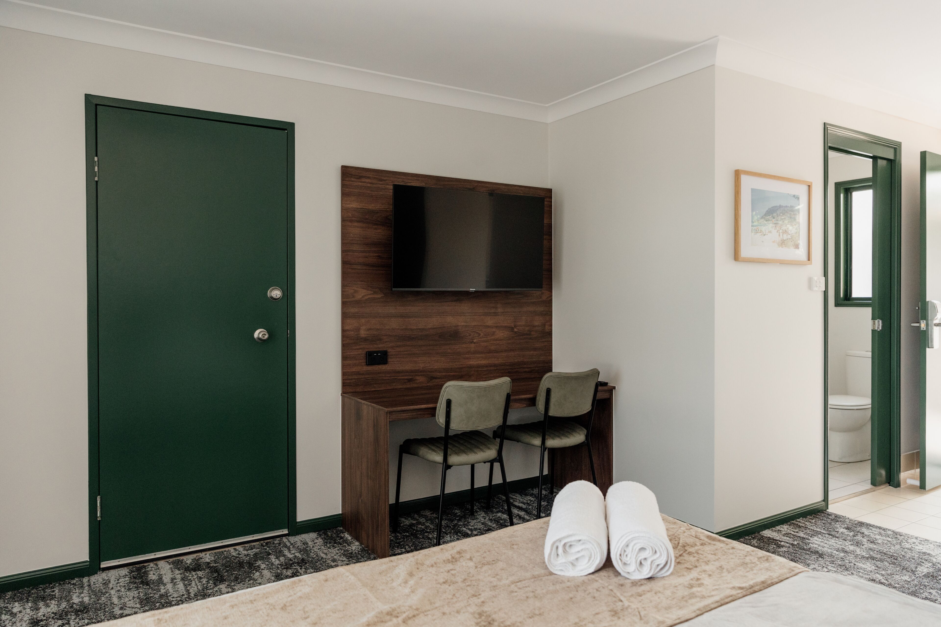 Habitación de hotel con puerta verde, televisor de pantalla plana montado en un panel de madera, dos sillas, cama con alfombra color crema y un cuadro en la pared.