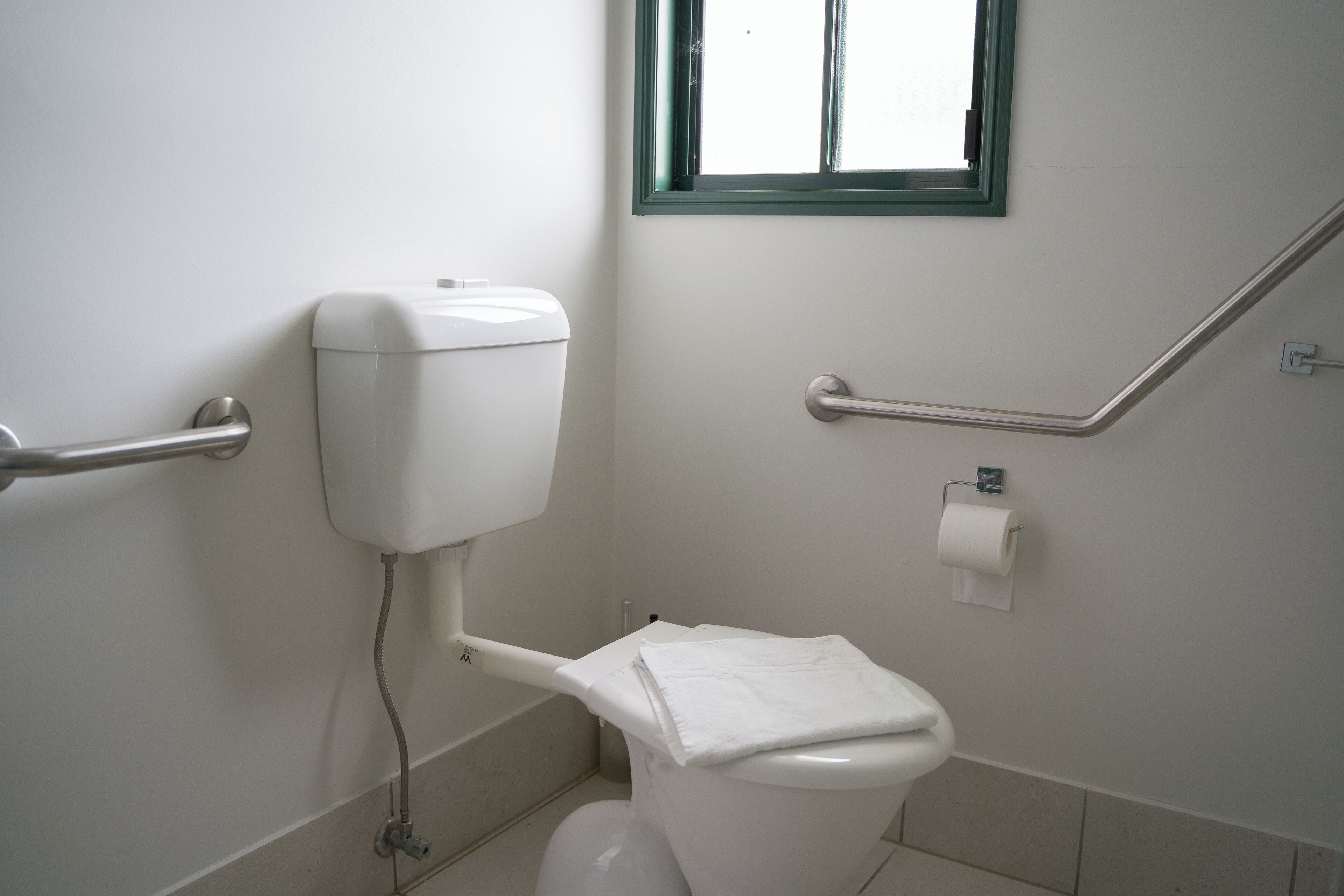 A white toilet with a towel on top, a handrail, and a window above it.