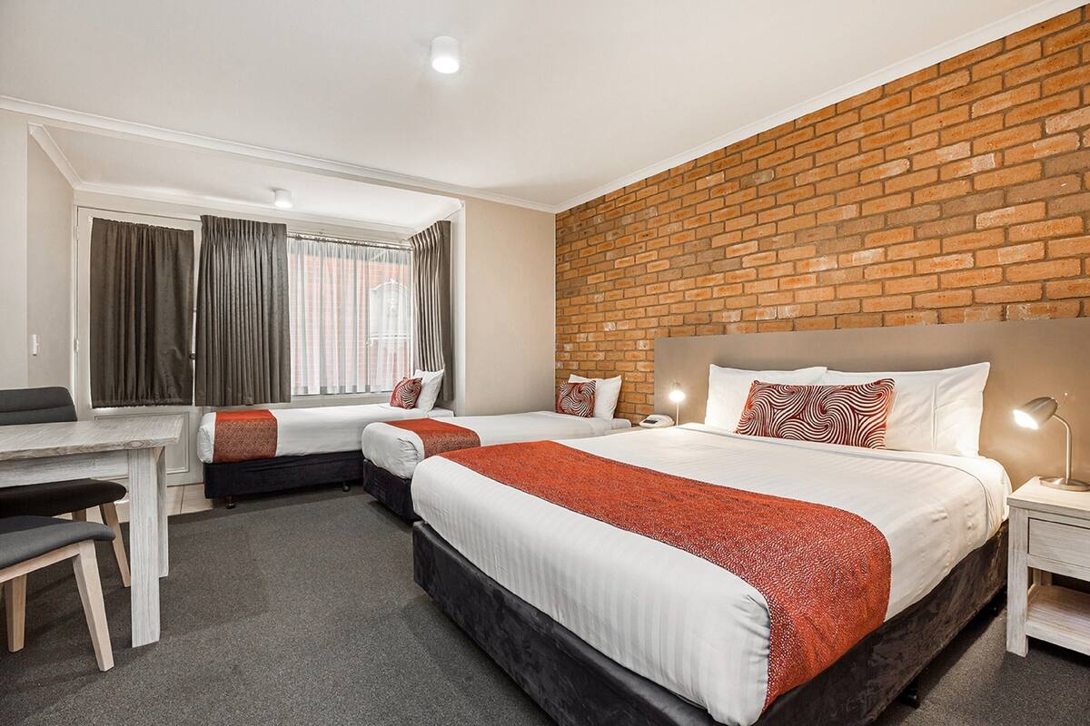 Quarto de hotel com duas camas, uma escrivaninha e uma parede de tijolos.