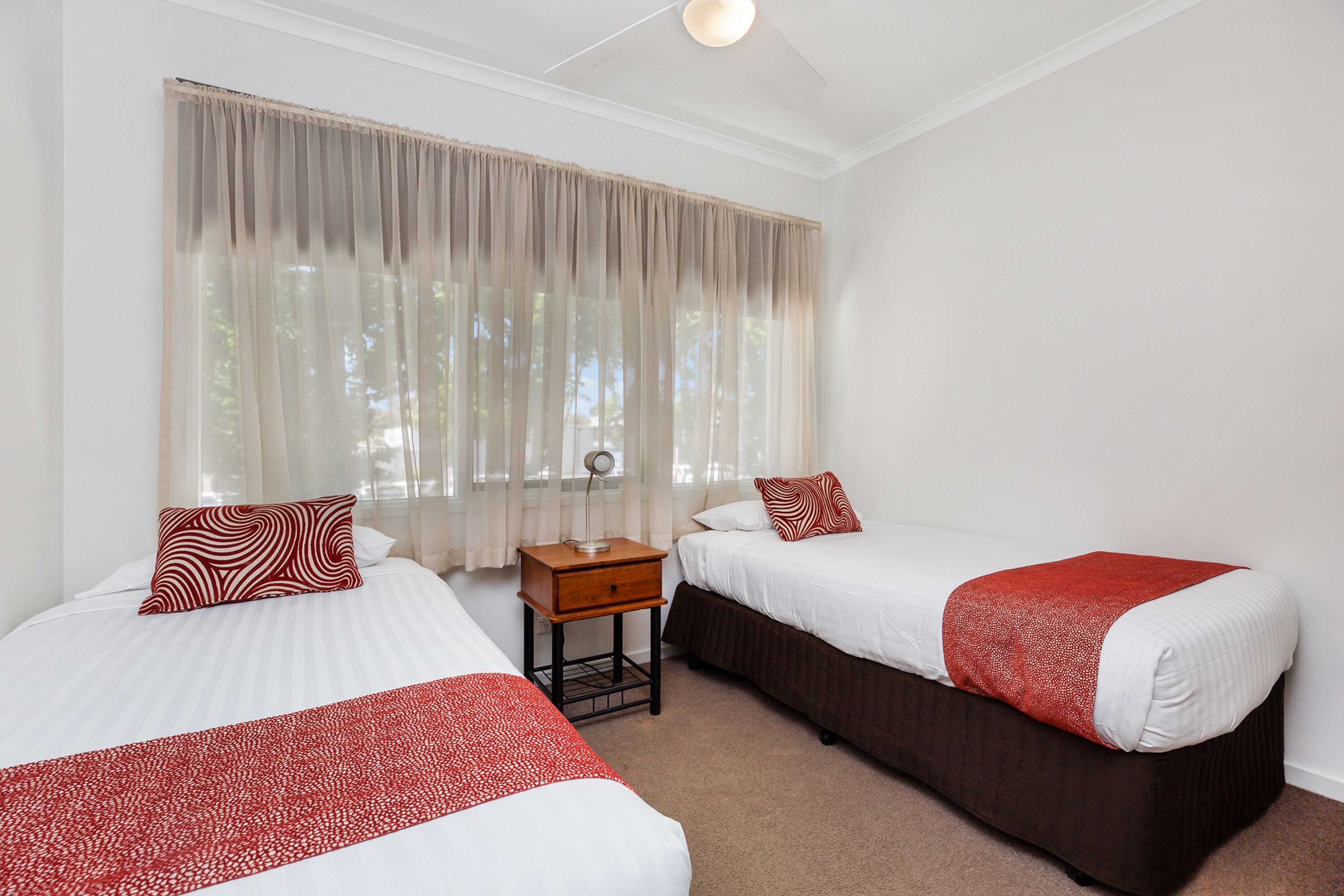 A hotel room with two beds, a nightstand, and a window with sheer curtains.