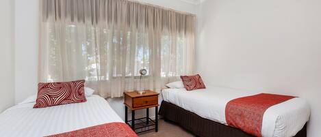 A hotel room with two beds, a nightstand, and a window with sheer curtains.