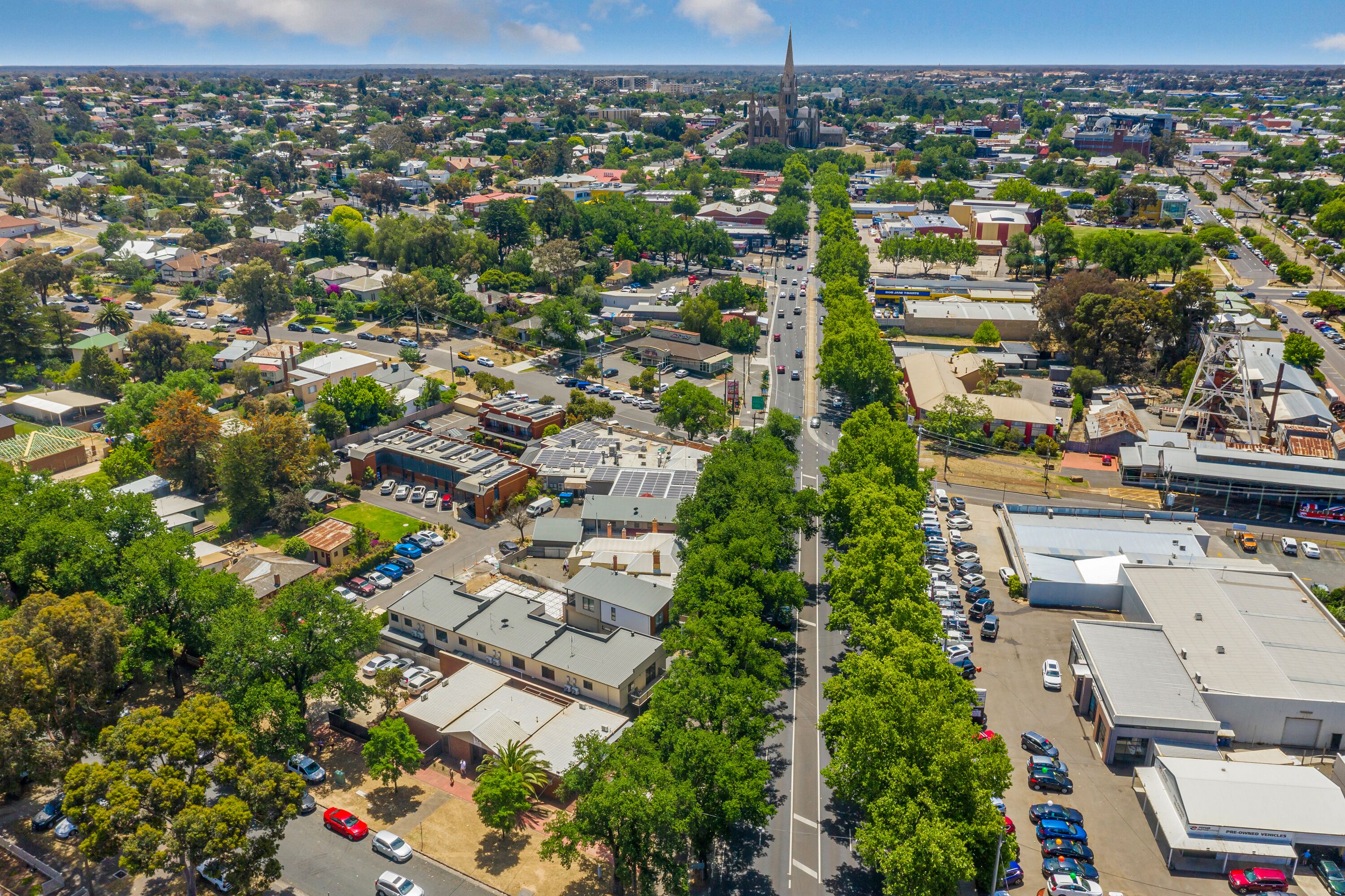 An aerial view of a town with a prominent church spire, a main road lined with trees, and various buildings including a large commercial structure.