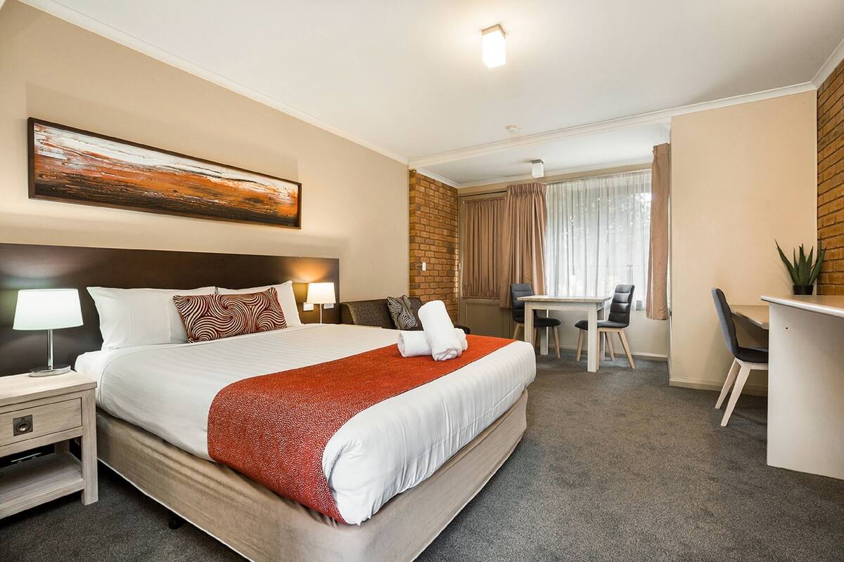 Quarto de hotel com uma cama grande, uma escrivaninha, uma cadeira, um abajur e um quadro na parede.