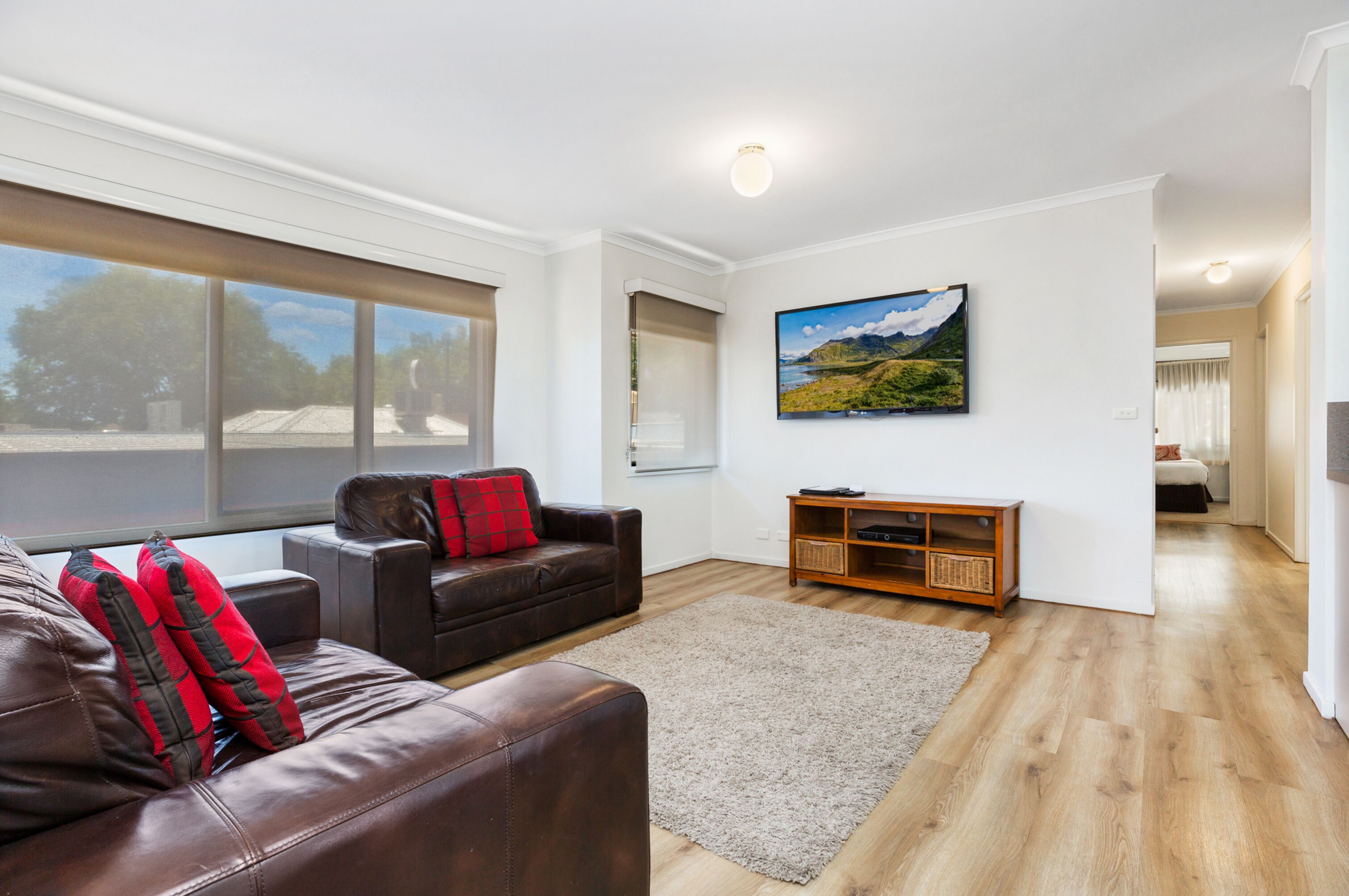 A modern living room with a brown leather sofa, a wooden entertainment unit, and a large window offering a view of the outdoors.