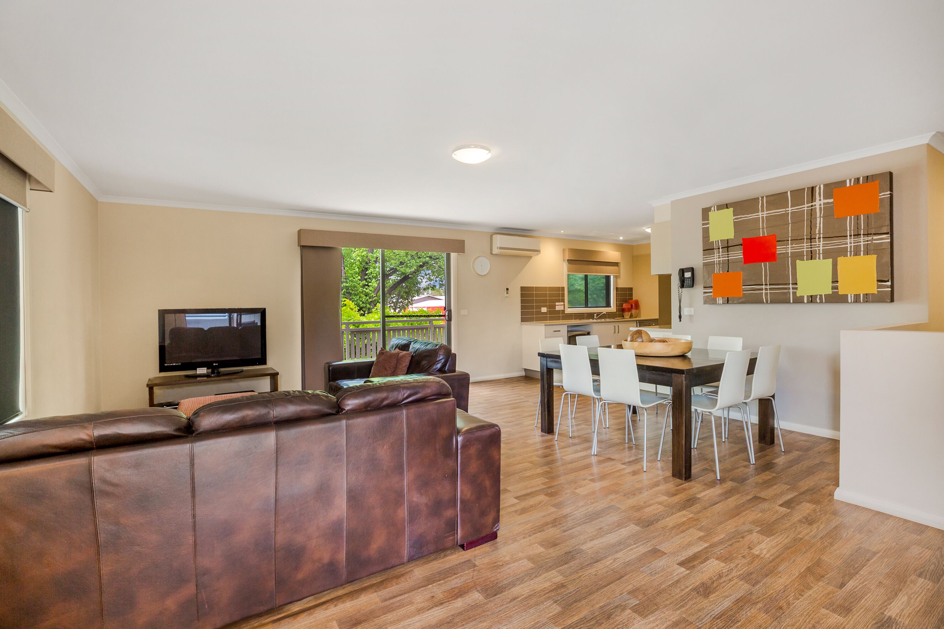 A modern living room with a brown leather sofa, a flat-screen TV, and a dining area with a wooden table and white chairs.