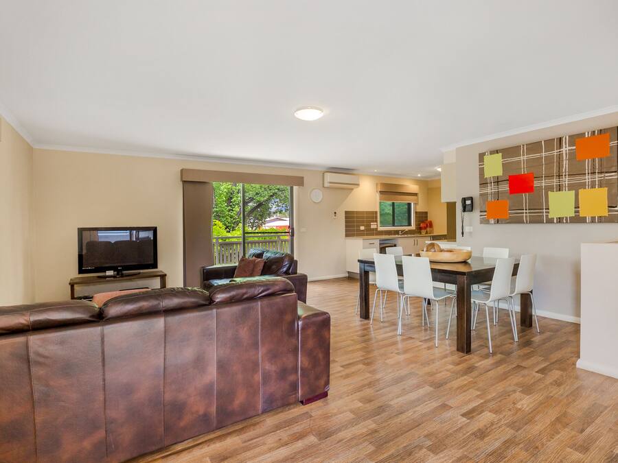 A modern living room with a brown leather sofa, a flat-screen TV, and a dining area with a wooden table and white chairs.