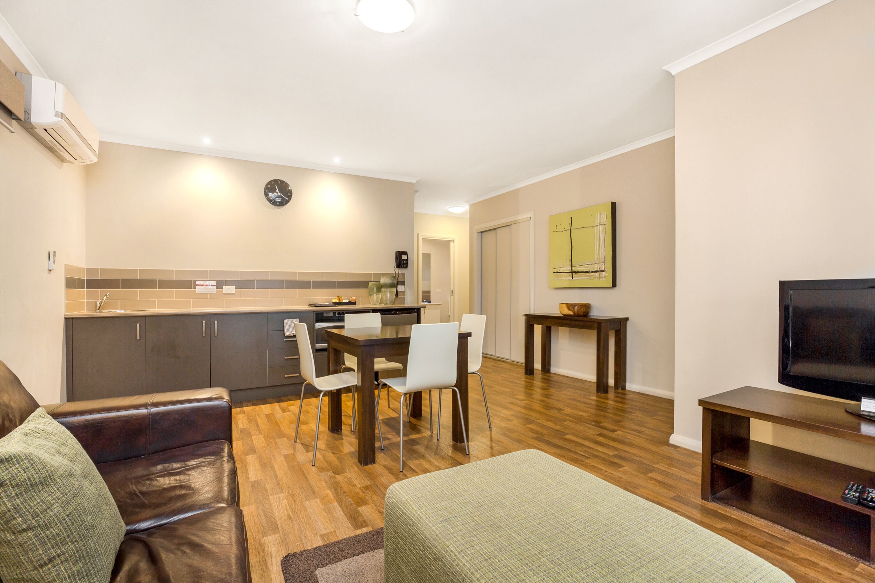 Una sala moderna con un sofá de cuero, una mesa de comedor con sillas, un mueble para la televisión y una zona de cocina con armarios y un reloj.