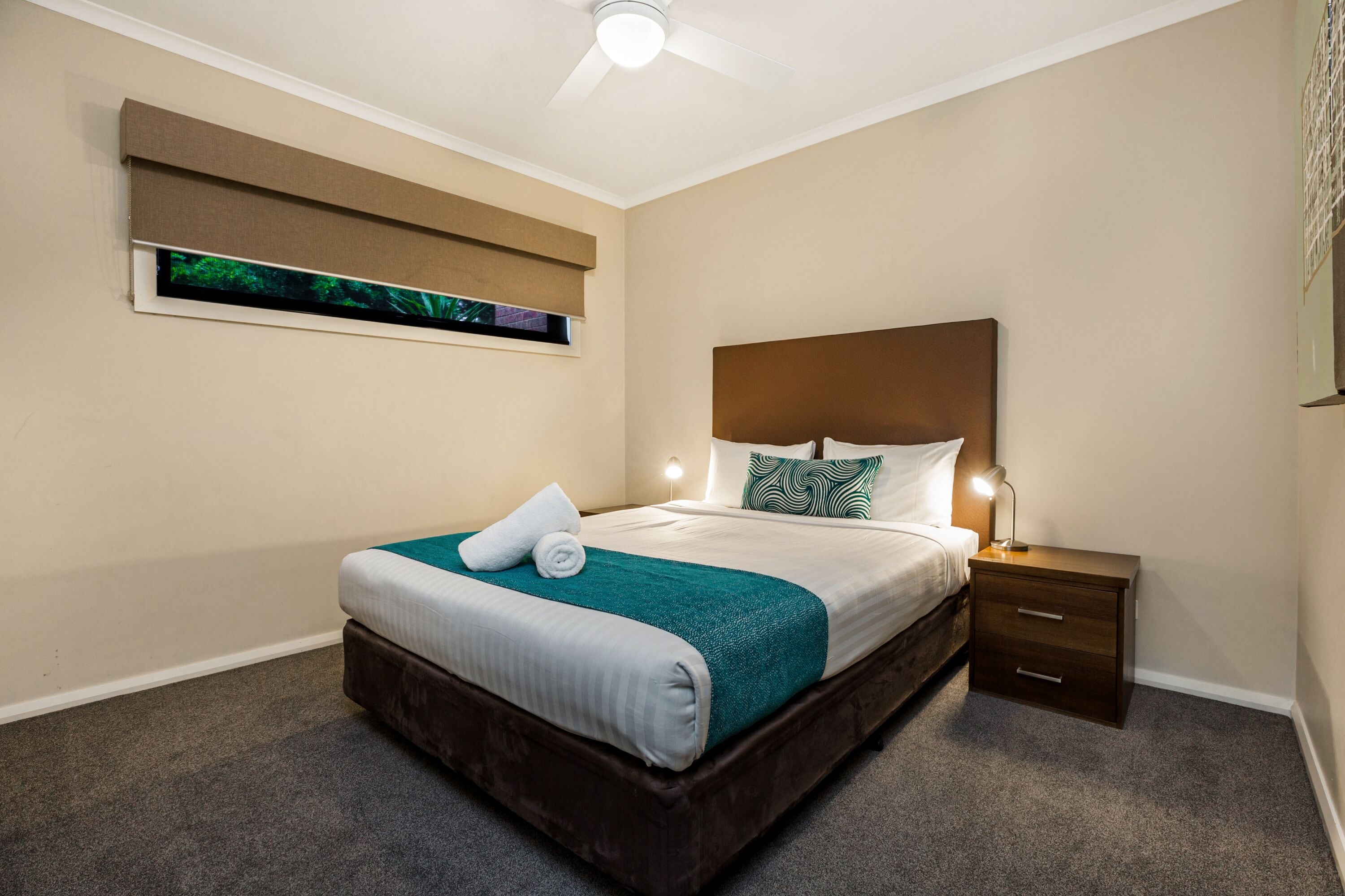 Un lit bien fait, agrémenté d’un plaid turquoise, une table de chevet avec une lampe et un ventilateur de plafond.