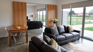 A modern living room with a sofa, a dining table, and a large window offering a view of the outdoors. - Relbia Lodge (Relbia)