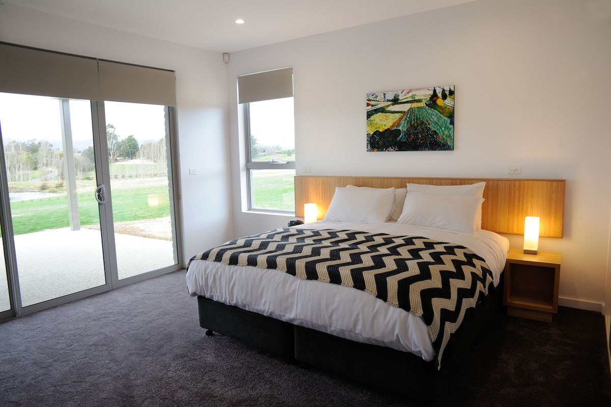 A bedroom with a large bed, two bedside tables with lamps, a sliding glass door leading to a balcony, and a painting on the wall.
