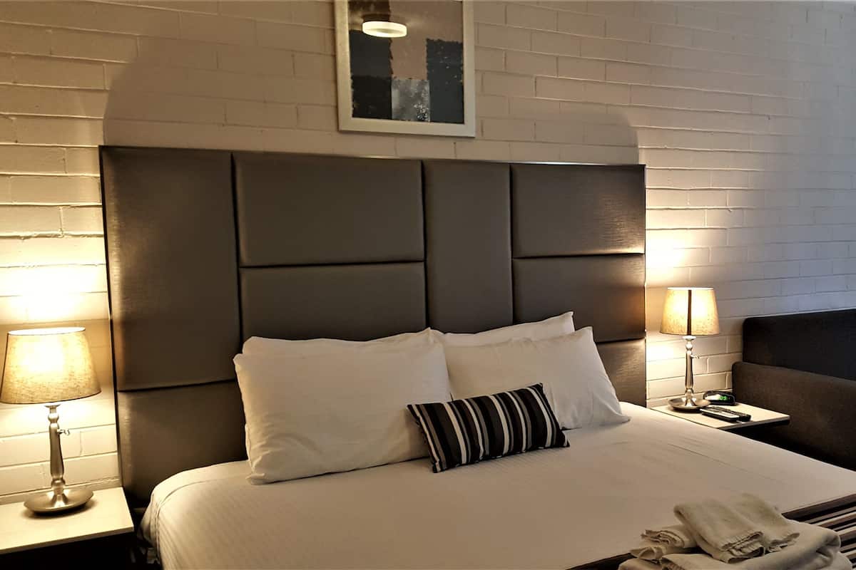 Quarto de hotel com uma cama grande, duas luminárias de cabeceira, uma cadeira e uma mesinha com um relógio.