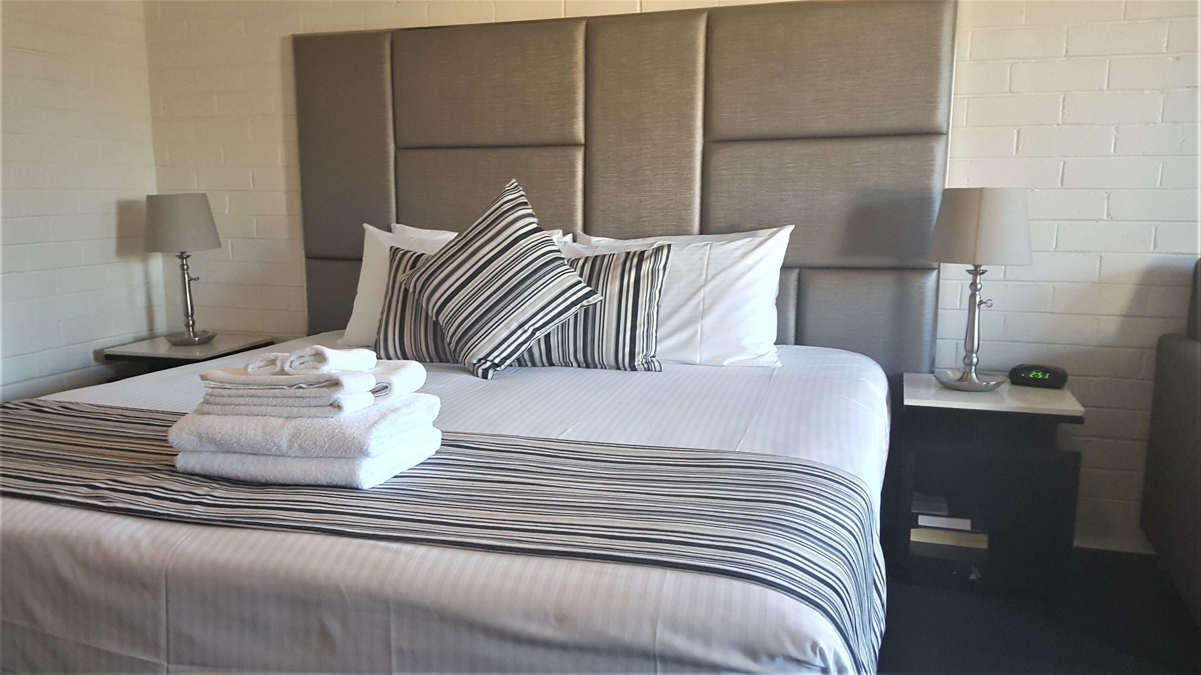 Una cama bien hecha con ropa de cama a rayas, una pila de toallas dobladas y almohadas a rayas. Hay dos mesitas de noche con lámparas y un reloj digital.