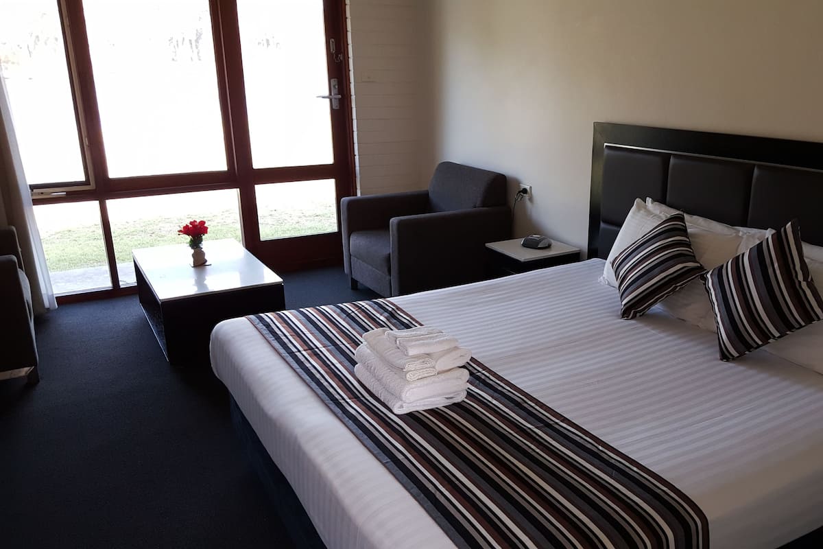 Quarto de hotel com uma cama grande, uma mesa de cabeceira, uma cadeira e vista para o exterior através de uma janela ampla.