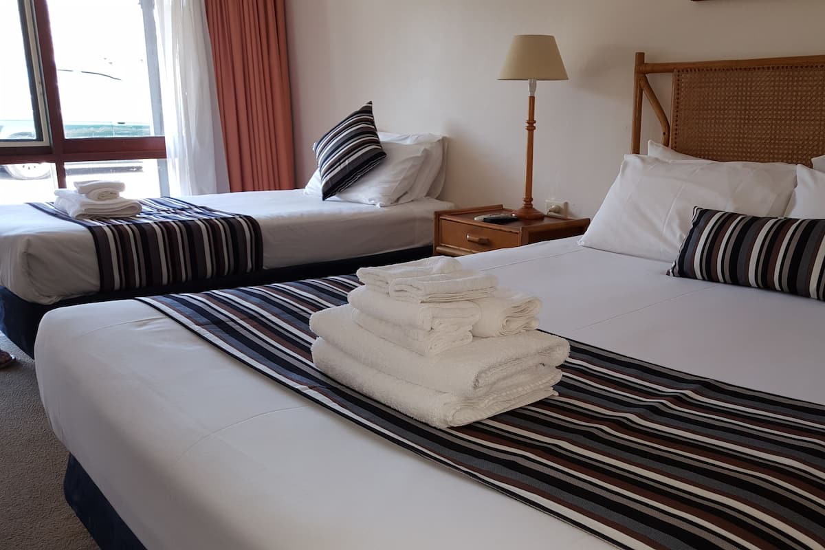 Quarto de hotel com duas camas, cada uma com roupa de cama e almofadas listradas. Há uma pilha de toalhas no chão.
