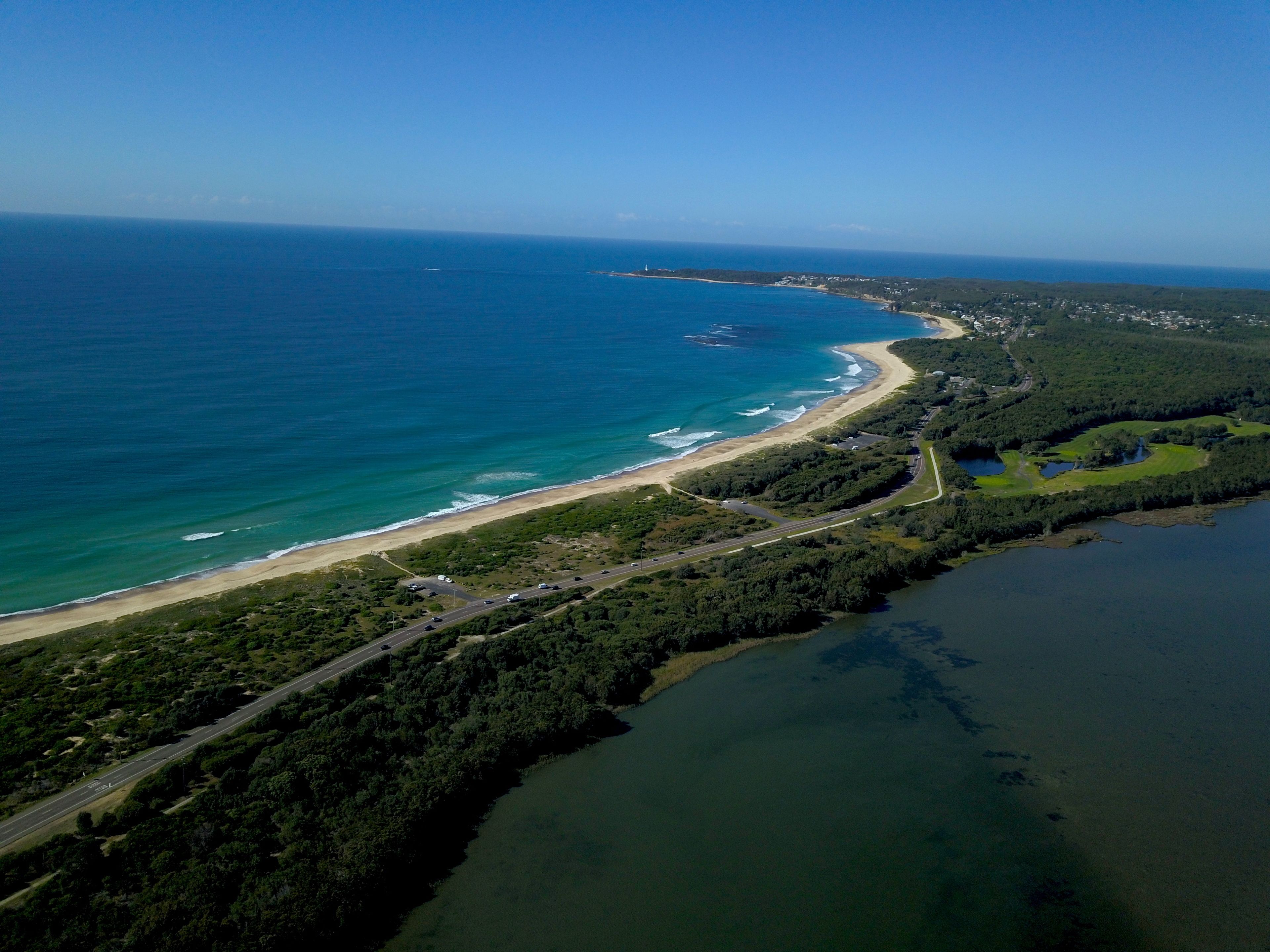 Vista aérea de una zona costera con playa, carretera y zona boscosa.