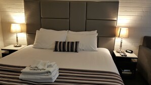 A neatly made bed with white linens and striped pillows, two bedside lamps, and a stack of folded towels on the bed. - Hibiscus Lakeside Motel (Budgewoi)