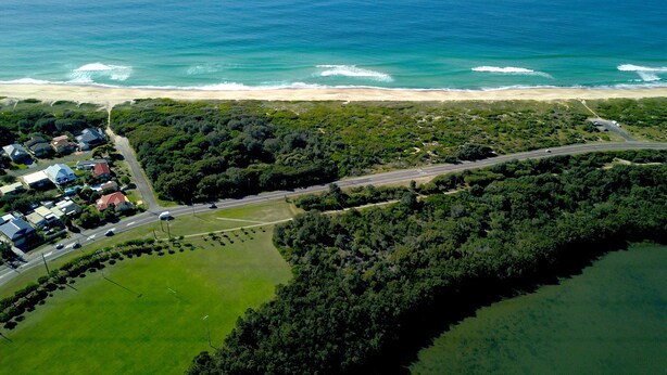 An aerial view of a coastal area with a beach, a road, and a residential area with houses. - Hibiscus Lakeside Motel (Budgewoi)