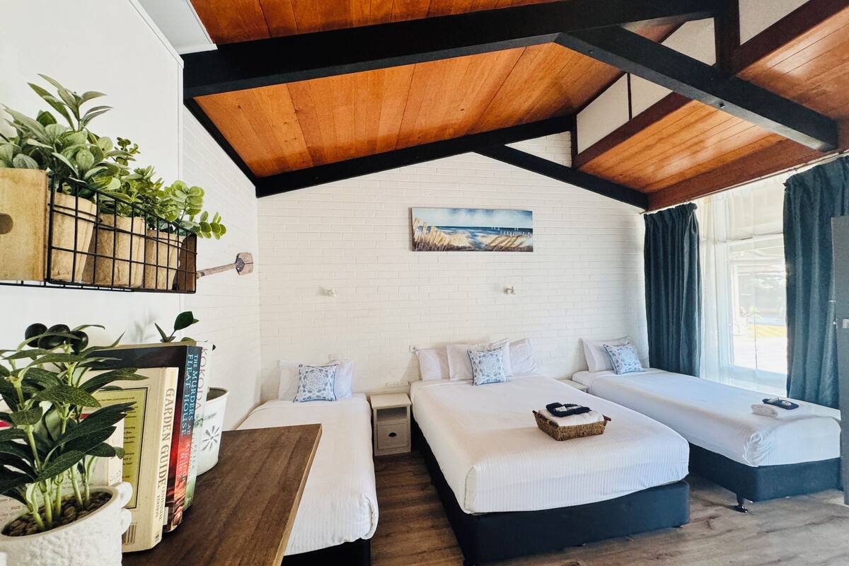 A cozy bedroom with a wooden ceiling, a white brick wall, and a large window with curtains. There are two beds with white bedding and a small table with a basket.