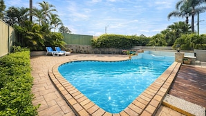 A swimming pool with a curved edge, surrounded by a brick deck, palm trees, and a green fence. - Jetty Motel The Entrance (Long Jetty)